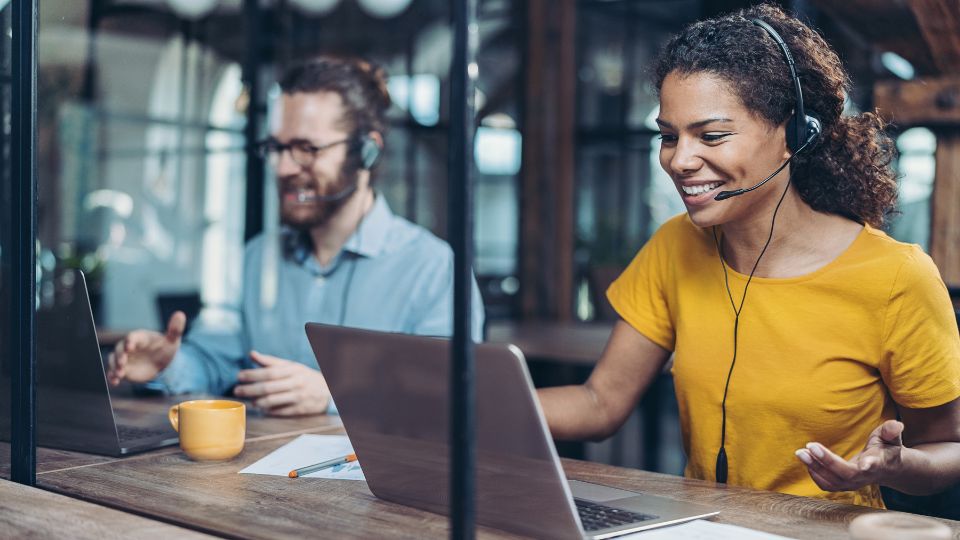 woman and man sitting at a co-working space with headsets on working on their laptops and happy dispositions.